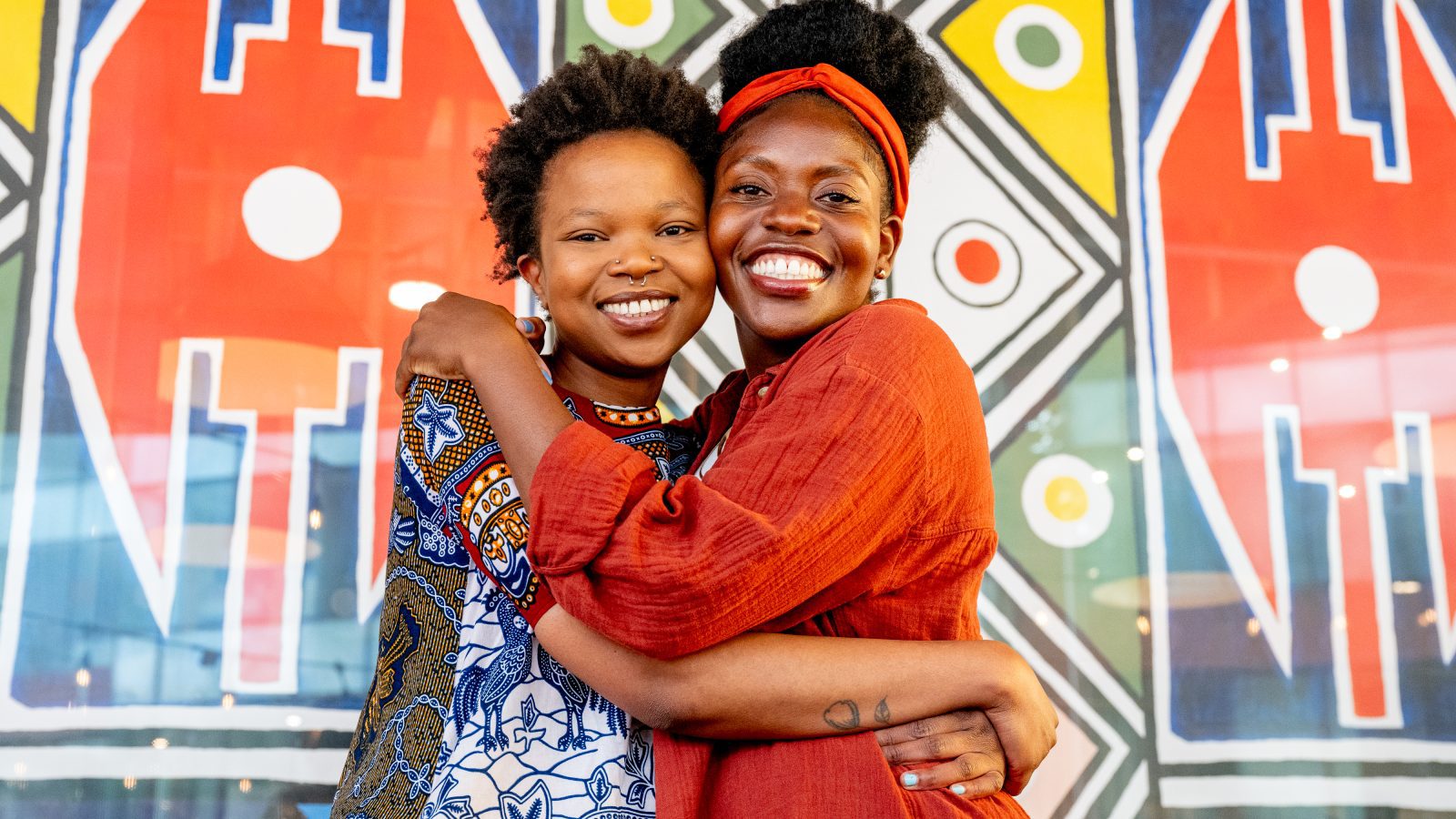 A non-binary black person and a black woman smiling in front of a colourful artwork at the Horizons Festival.
